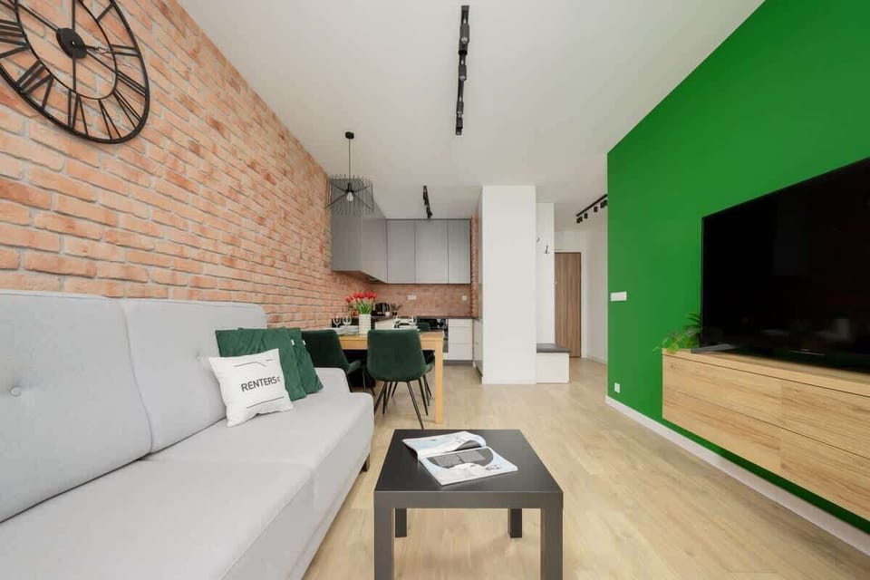 A living area with a wall clock, gray couch, and stylish decor against green and brick feature walls.