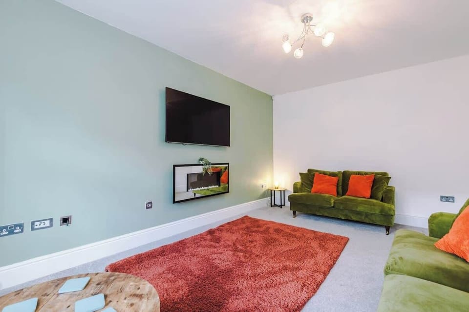 A bright, contemporary living space with plush green sofas, warm orange cushions, and a soft rug — designed for ultimate relaxation and cosy evenings.