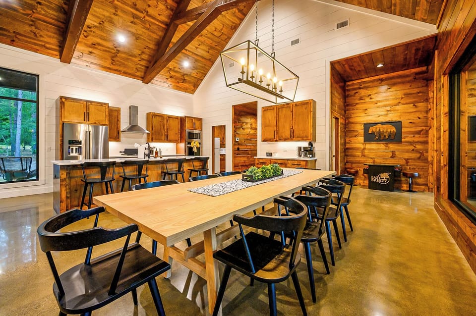 Modern luxury kitchen with vaulted wood ceilings, stainless appliances, and a massive light-wood dining table with black chairs.