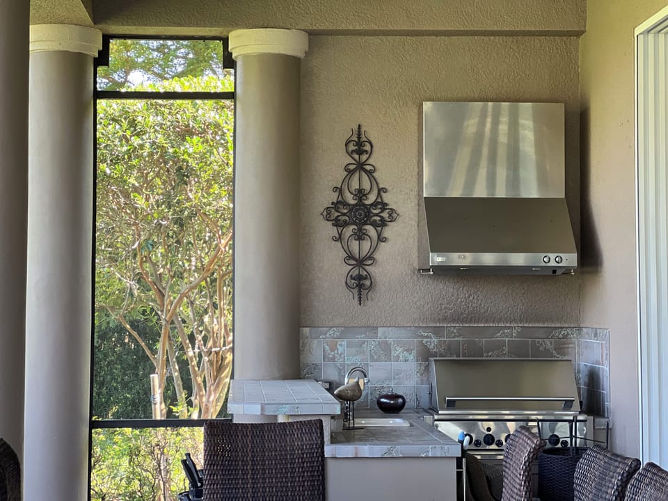 Outdoor kitchen with a stainless steel grill, tiled countertop, and decorative wall art. Two wicker chairs are partially visible. The area is enclosed by pillars and screens, with trees outside.