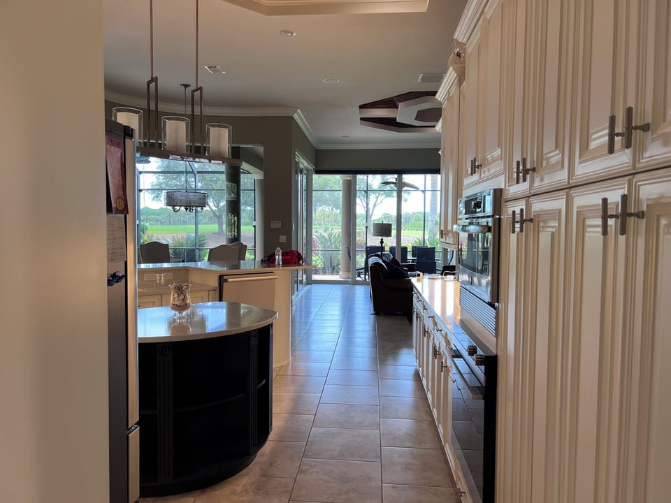 A modern kitchen with white cabinetry and an island granite countertop leads to an open living area featuring large windows and a view of the outdoor greenery.