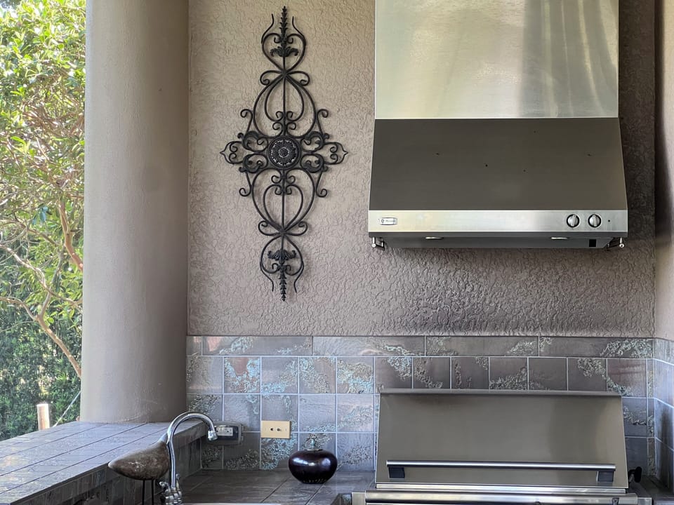 Outdoor kitchen area with a wall-mounted ventilation hood, a decorative wrought iron piece, a granite countertop, small sink, and tiled backsplash. Trees and greenery are visible in the background.
