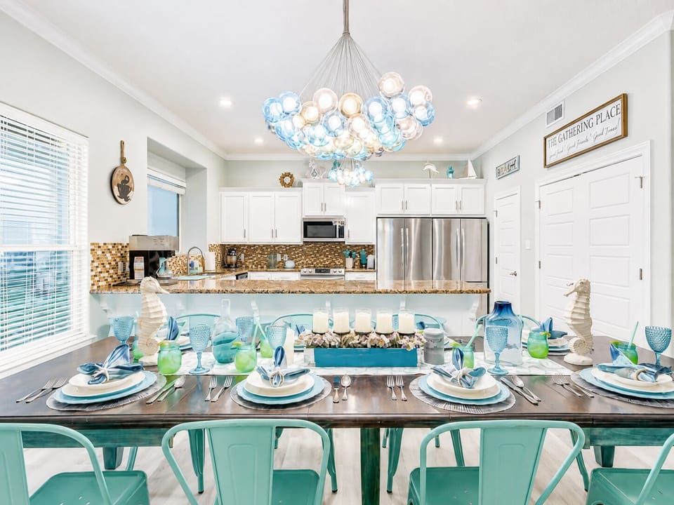 Coastal dining room with a large table, cushioned bench seating and a modern chandelier, offering a welcoming space for shared meals and celebrations.