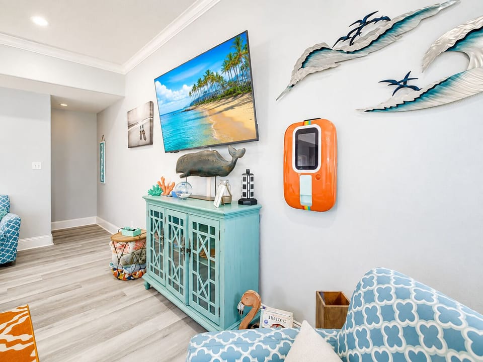 Coastal media area with a wall-mounted Smart TV, decorative console, accent chair and colorful beach-inspired artwork for laid-back indoor entertainment.