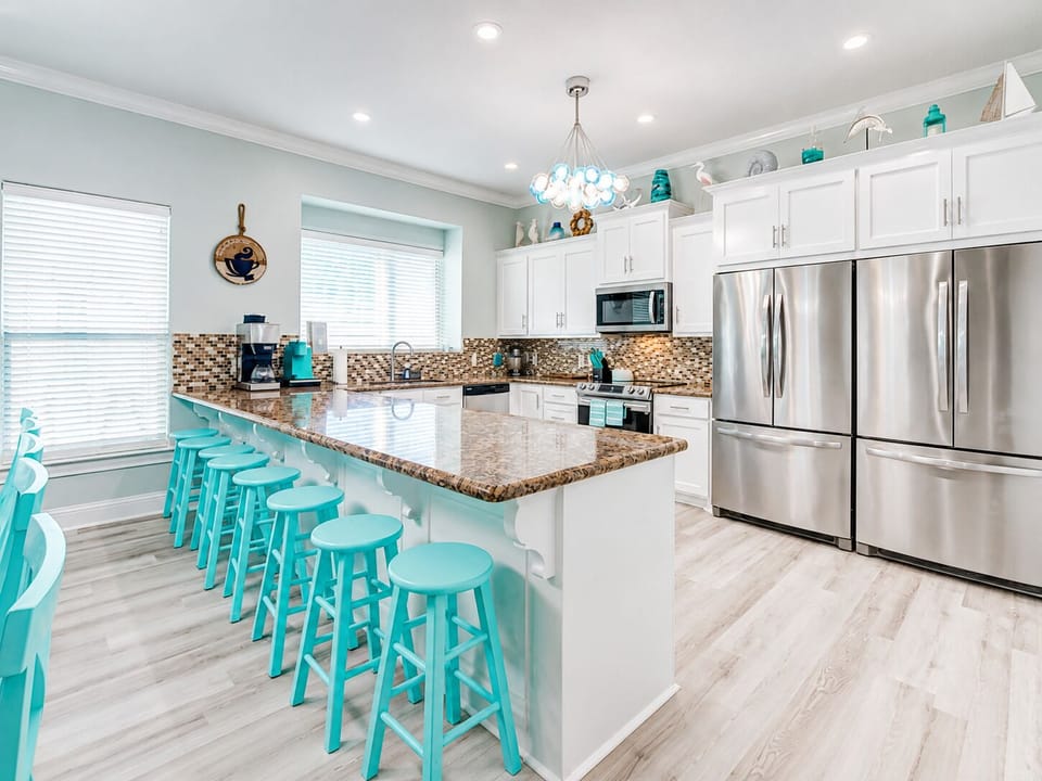 Fully equipped kitchen with stainless steel refrigerator, expansive granite counters, bright white cabinetry and a large breakfast bar with turquoise stools.