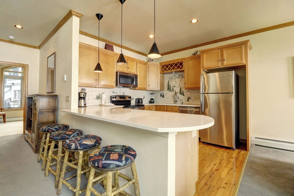 A modern kitchen with light wood cabinetry, stainless steel appliances, and a curved countertop with four patterned stools. There are hanging pendant lights above the counter and a backsplash featuring a scenic design.