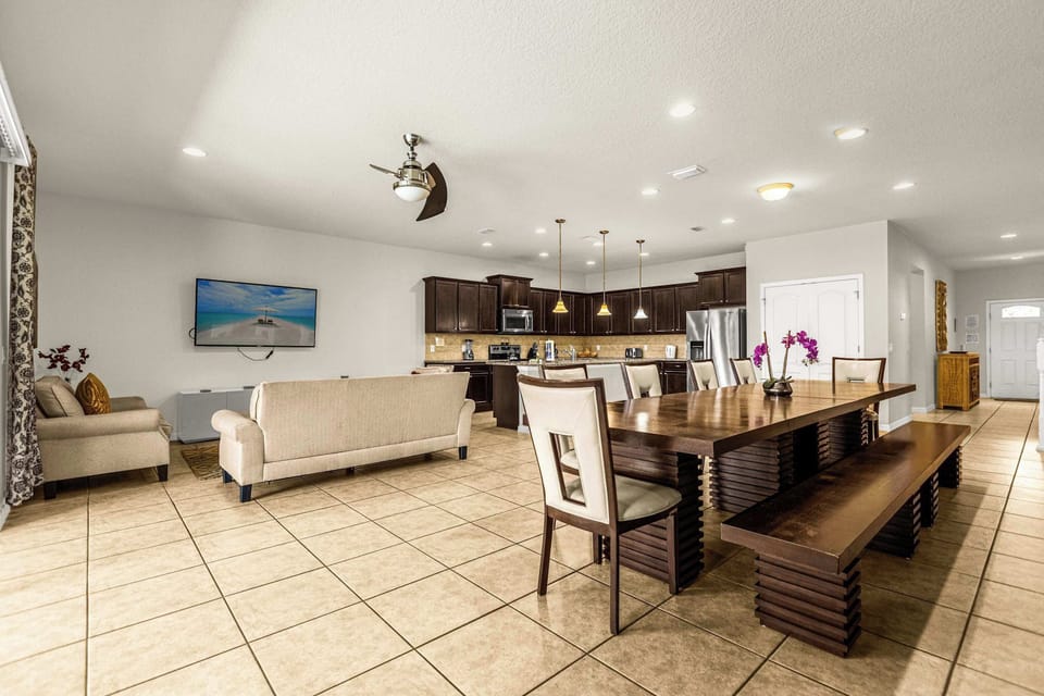 Open-plan living and dining area with a fully equipped dark-wood kitchen, beach-view TV screen, and long dining table for shared meals.