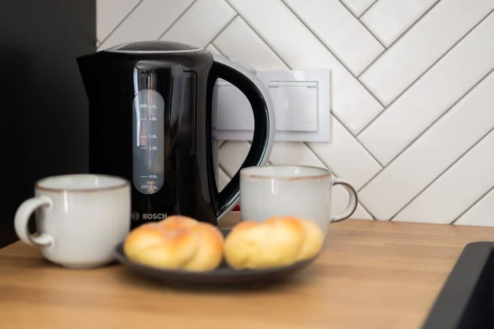 A close-up of a black electric kettle with two apples on a plate and a cup, set against a white tiled wall, creating a cozy breakfast nook.