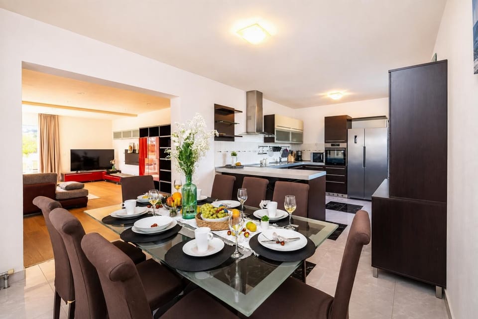 A modern open-plan dining and kitchen area features a glass-top dining table set for six with brown upholstered chairs, opening up to a contemporary kitchen with dark wood cabinets and stainless steel appliances.