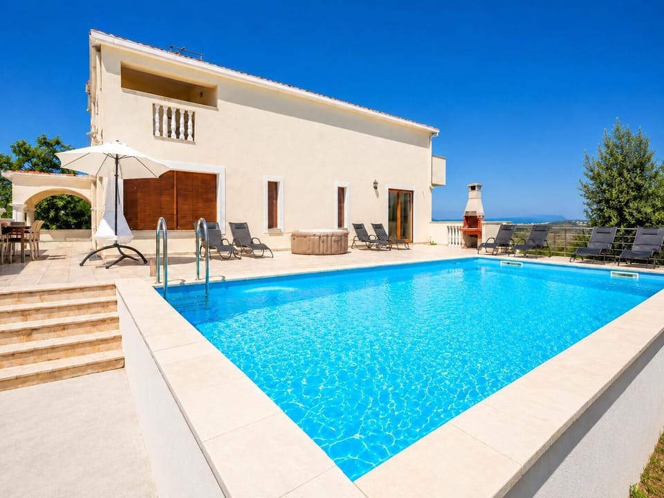This modern cream-colored villa features a large, crystal-blue swimming pool and a spacious stone patio lined with lounge chairs, a hot tub, and an outdoor barbecue under a clear blue sky.