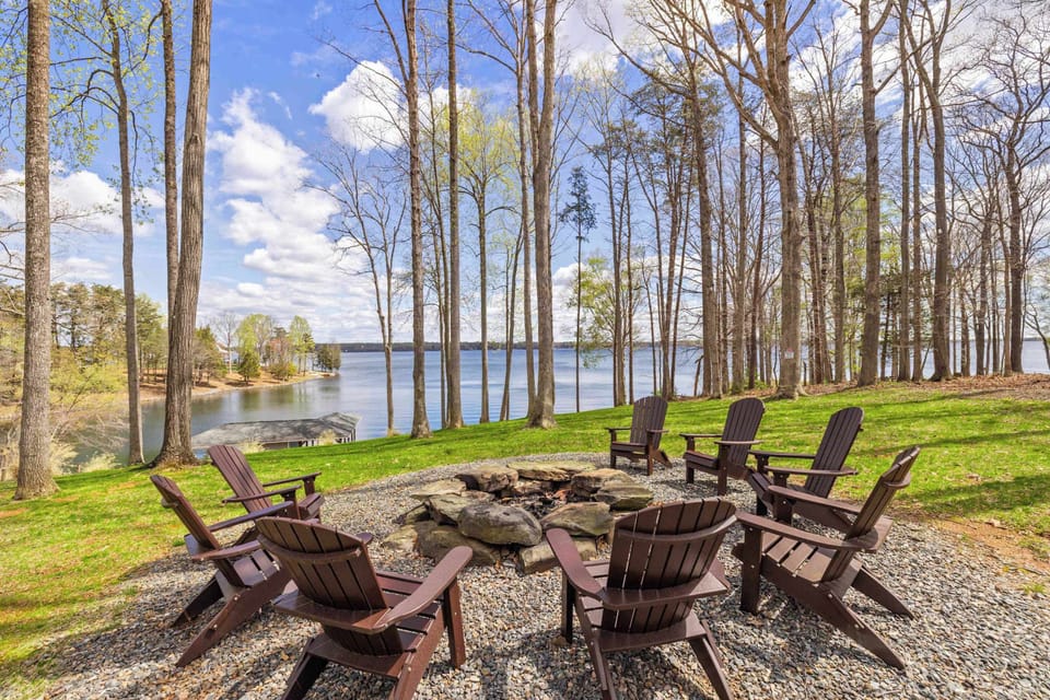 A circle of Adirondack chairs surrounds a stone fire pit, poised for lakeside relaxation amidst towering trees and a clear blue sky.