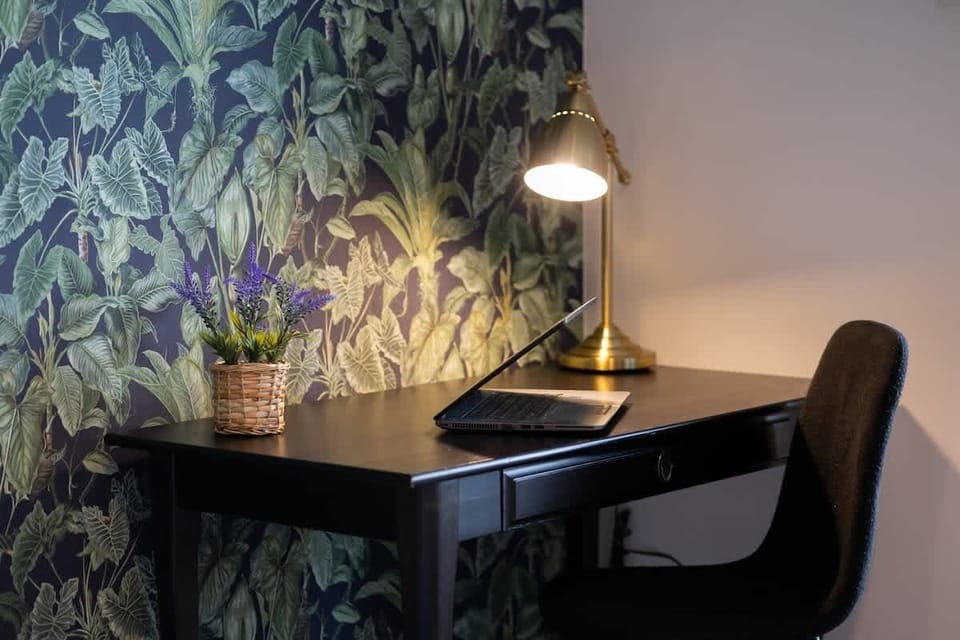A close-up of a desk setup in a corner of a room with green leafy wallpaper. The black chair and lamp create a cozy work nook.
