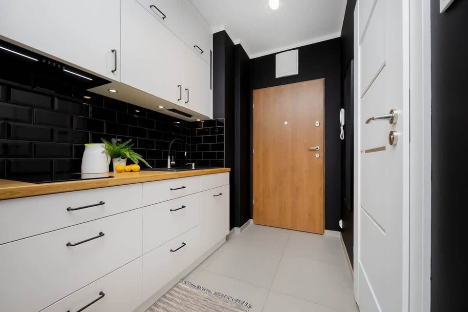 A narrow kitchen hallway with modern white cabinets and a black backsplash. A wooden door at the end adds contrast to the sleek design.