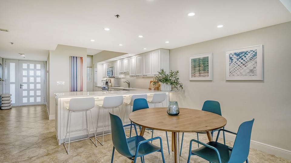 Dining area with round wood table, teal chairs, and white bar stools along a white kitchen island.
