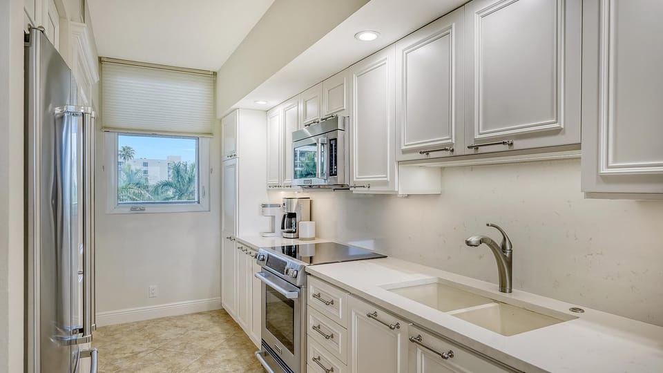 Galley kitchen with white cabinetry, stainless steel appliances, marble countertops, and a window with palm tree views.