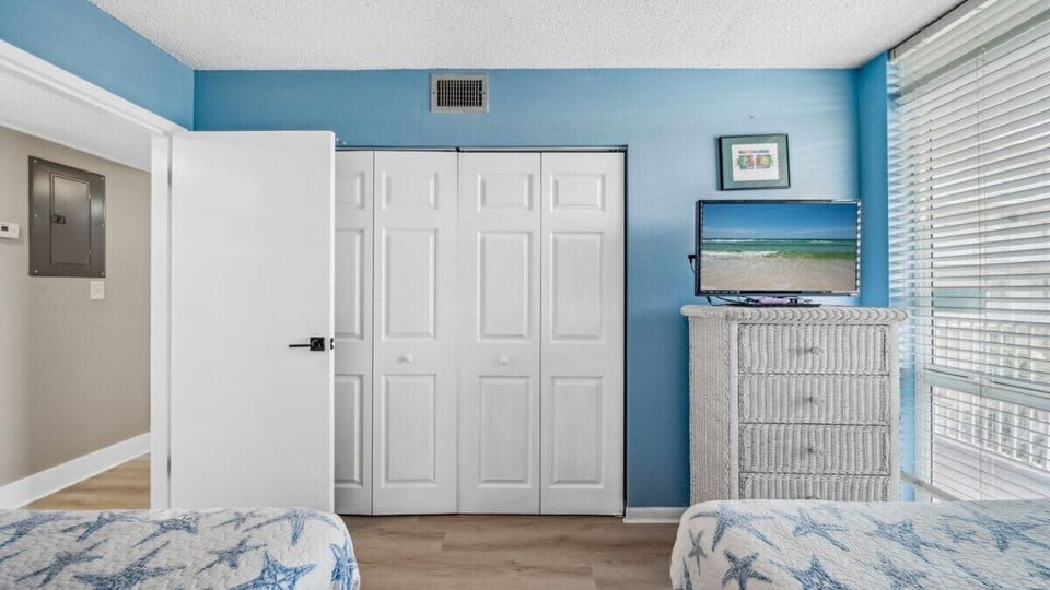 Guest room with full closet, wicker dresser, TV, and bright blue walls—fun, coastal, and full of natural light.
