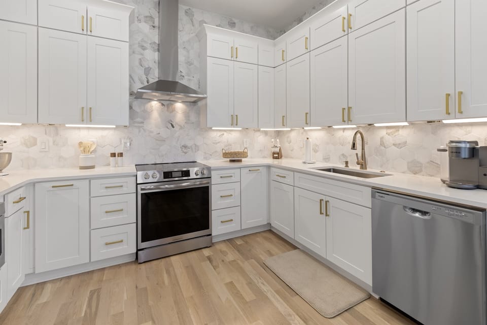 Step into this bright, modern kitchen with pristine white cabinets, elegant gold hardware, and gleaming marble backsplash that creates your perfect culinary haven.
