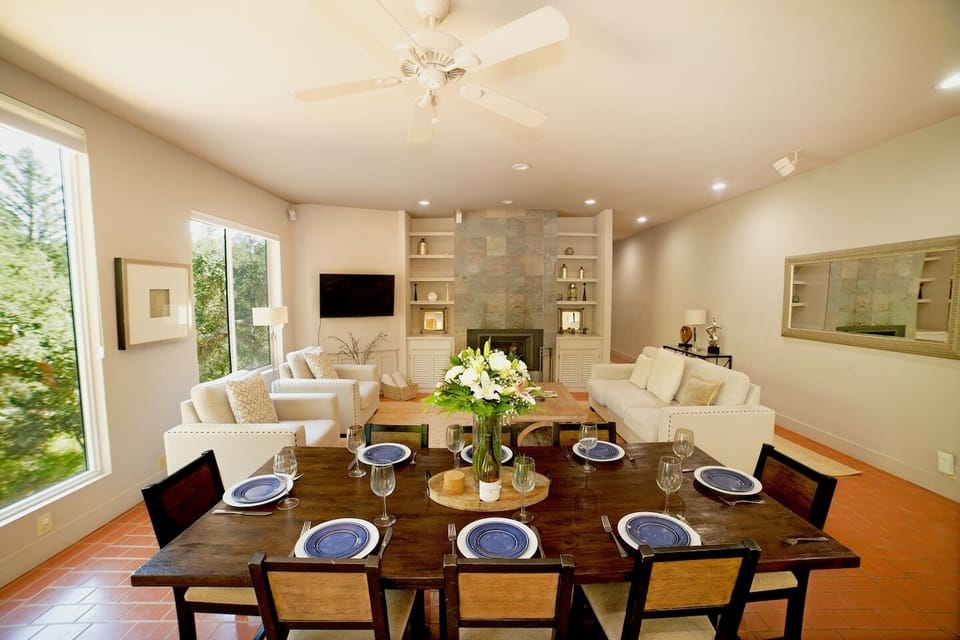Elegant open-plan dining and living area with a dark wood table set for six, white sofas, stone fireplace with built-in shelves, mounted TV, and large windows framing lush garden views. Terracotta floors and floral centerpiece add warmth.