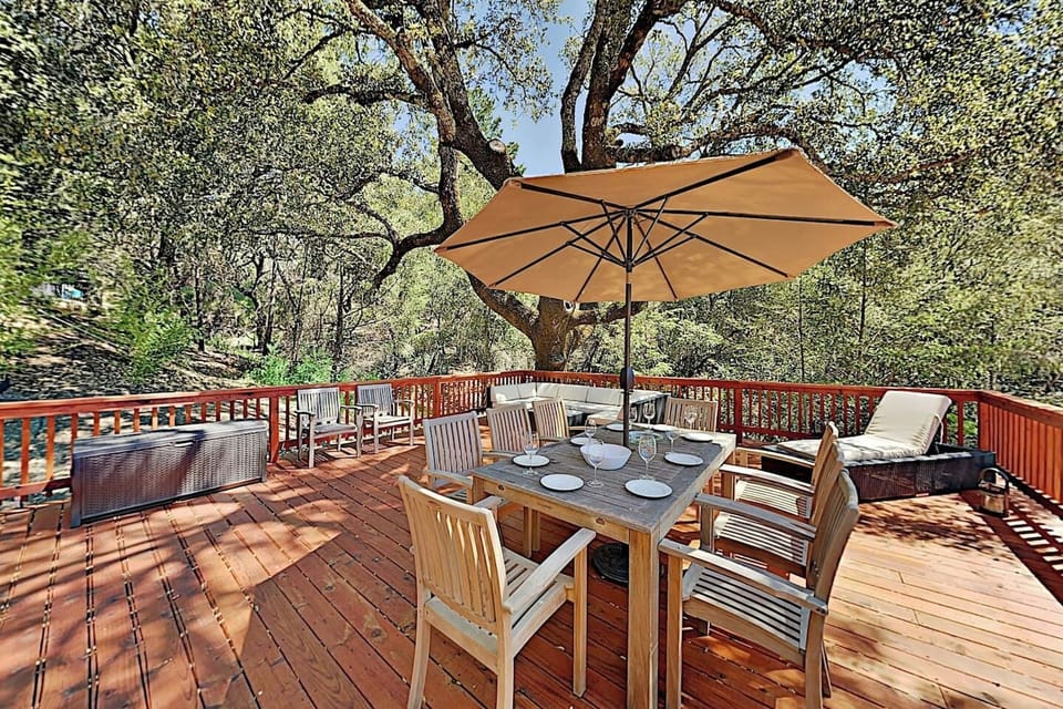 Sun-dappled wooden deck shaded by mature oak trees, featuring a dining table for six under a tan umbrella, plus lounge seating and a fire pit area. Surrounded by woodland scenery, perfect for outdoor dining and relaxation.