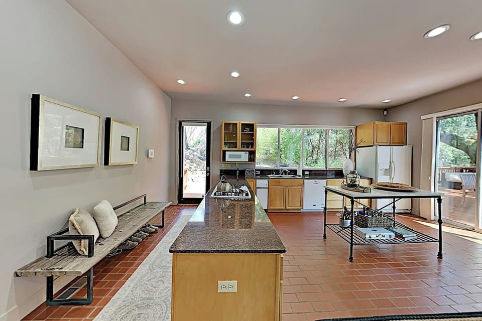 Bright, open kitchen with granite island, oak cabinets, terracotta tile floors, and large windows with garden views. Features a gas cooktop, white appliances, and a rustic bench seating area, blending function with a relaxed, airy feel