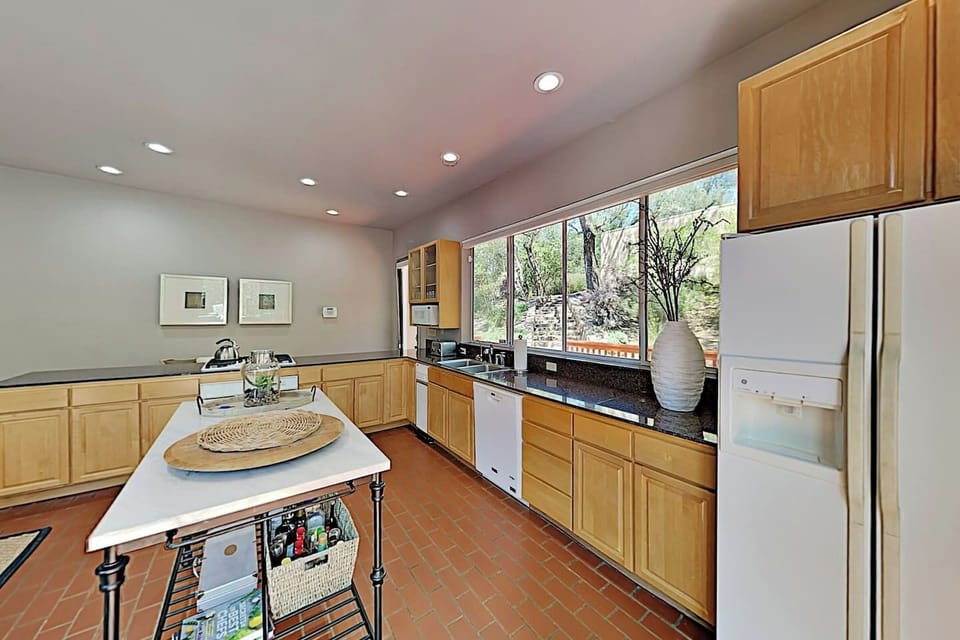 Well-equipped kitchen with honey oak cabinets, dark granite countertops, terracotta tile floors, and a large window overlooking the wooded garden. Features a freestanding island, white appliances, and recessed lighting throughout