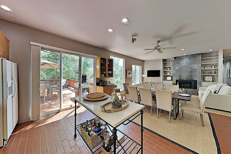 Open-plan kitchen, dining, and living area with terracotta floors, stone fireplace, built-in shelves, and mounted TV. Large sliding doors open to a sunny deck with outdoor dining, seamlessly blending indoor and outdoor living