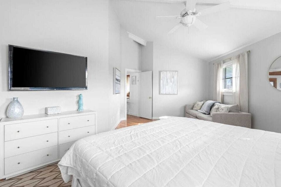 Relax in your serene bedroom with plush bedding, modern TV, and cozy seating area bathed in natural light.