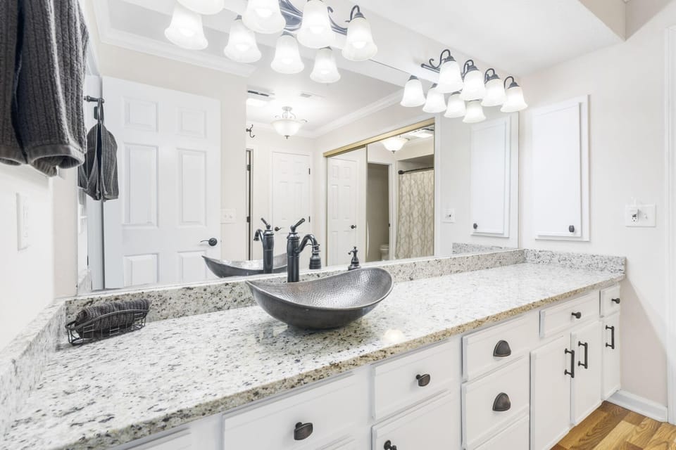Bathroom countertop stretches with speckled granite, white cabinetry, hammered metal vessel sink, and black faucet fixtures.