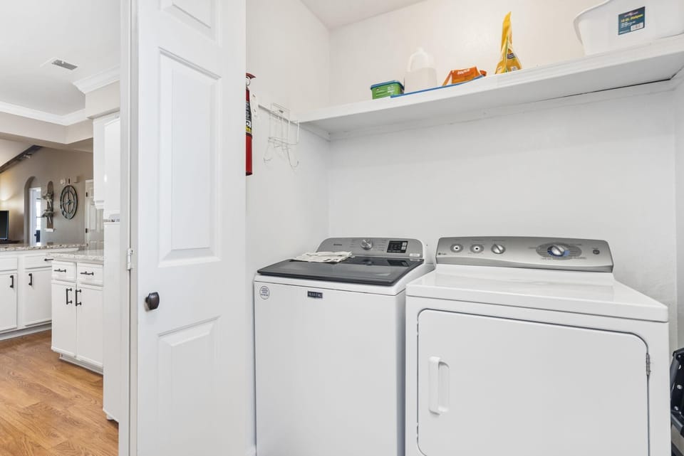 Laundry area with white Maytag washer and dryer, open shelf above stocked with detergent and supplies.