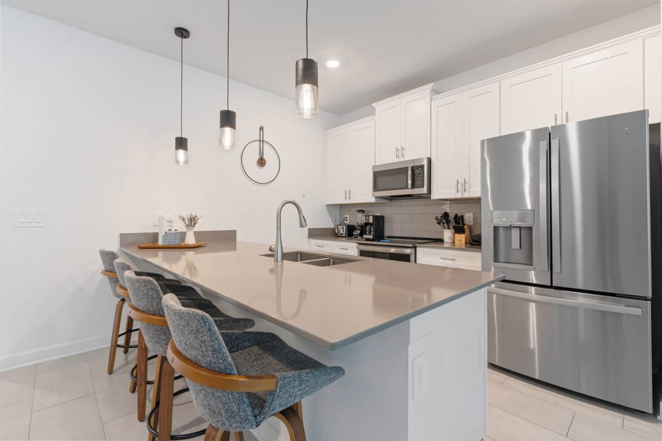This bright kitchen boasts stainless steel appliances and a spacious island with chic bar stools, ideal for casual dining. Modern pendant lighting highlights the clean, white cabinetry, inviting you to enjoy culinary adventures.