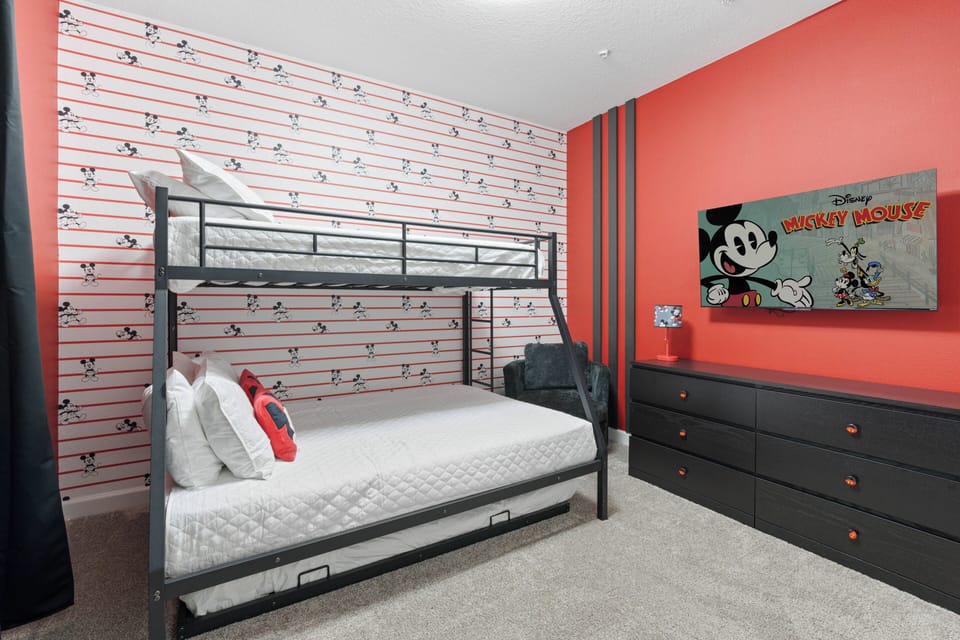 This vibrant bedroom boasts a fun red accent wall with a classic Mickey Mouse theme, making it perfect for guests. The sturdy metal bunk bed provides plenty of sleeping space, complemented by cozy linens and a stylish dresser.