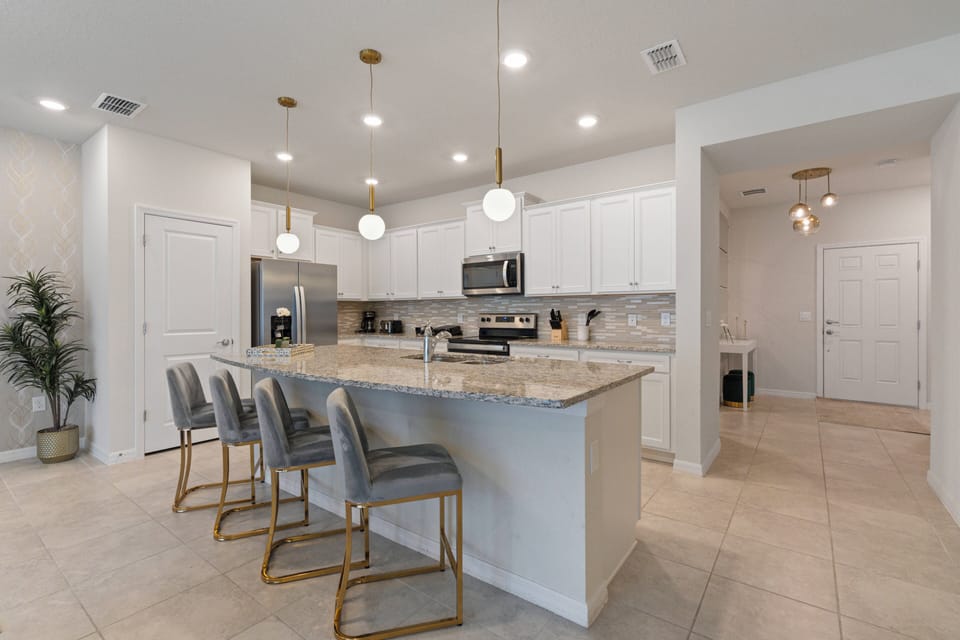This bright kitchen boasts a spacious island with sleek granite countertops and elegant gold-accented bar stools. Clean white cabinetry enhances the modern look, while pendant lights bring a warm touch to the inviting space.