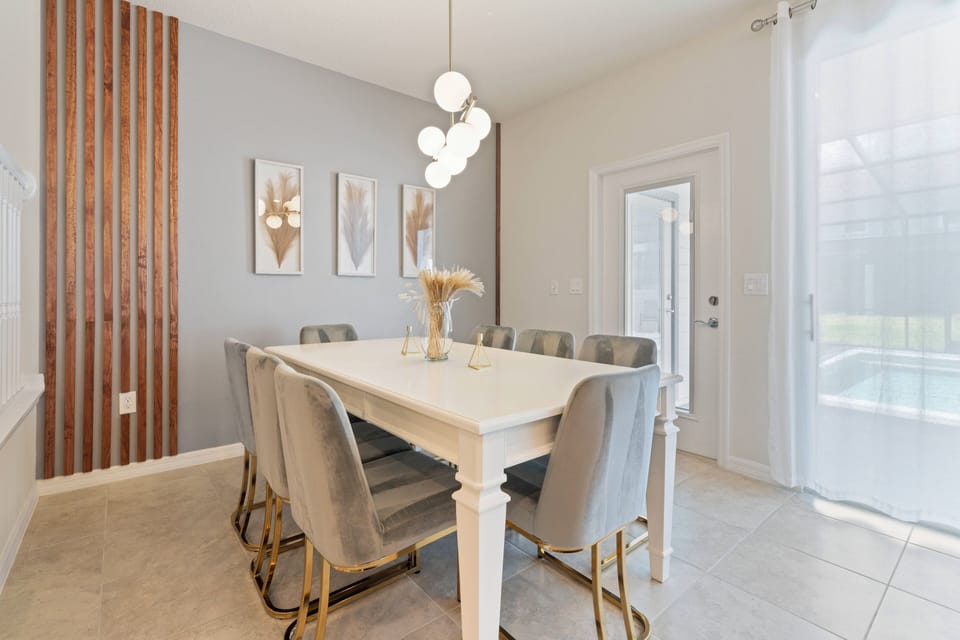 This bright dining area features a chic white table and plush gray chairs, perfect for shared meals. Soft natural light through sheer curtains creates an inviting atmosphere, beautifully enhanced by stylish wall art.
