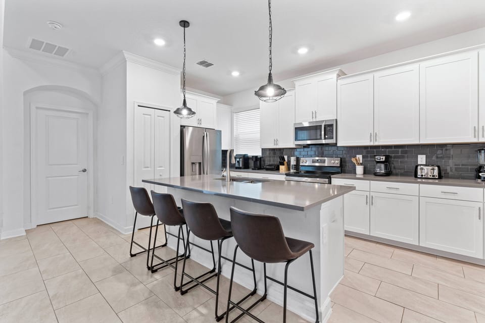 This modern kitchen boasts a spacious island with stylish bar stools, ideal for casual dining or your morning coffee. The white cabinetry and dark backsplash create a bright, inviting space for all your culinary adventures.