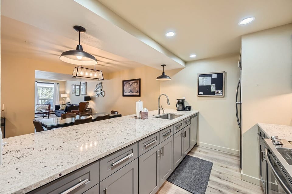 A modern kitchen with gray cabinets, white granite countertops, pendant lighting, a sink, and stainless steel appliances. The kitchen opens to a dining area and living room with hardwood flooring.