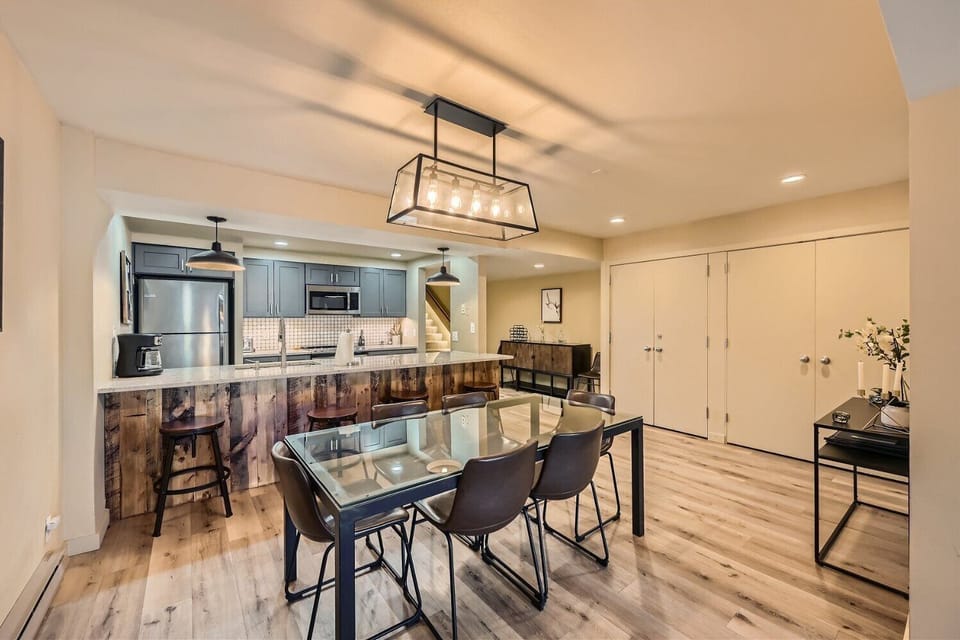 Modern kitchen and dining area with a glass-top dining table, four chairs, a two-tone kitchen with pendant lighting, a wooden island, and light hardwood flooring.