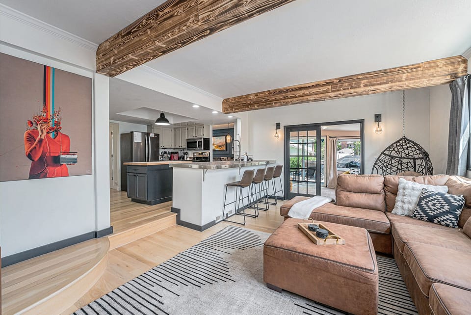 - Plush sectional sofa beneath rustic wood ceiling beams & natural light
- Fully equipped kitchen with breakfast bar seating, stainless steel appliances, & bold cabinetry
- Artistic wall features & a hanging chair add character to this stylish social hub