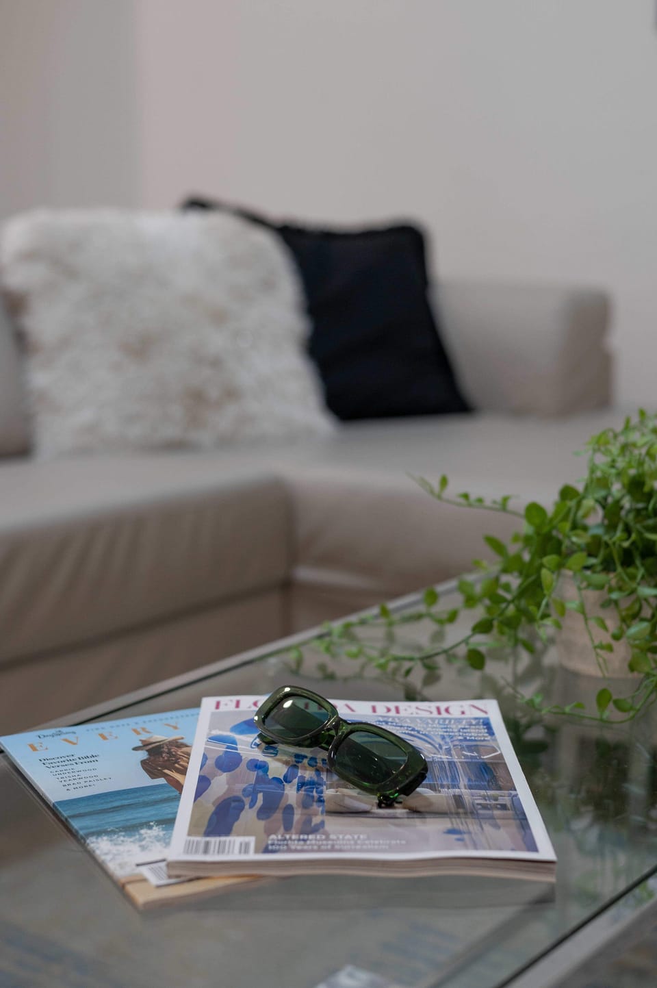 A cozy living area with a beige sofa, textured pillows, and a glass coffee table featuring magazines and sunglasses.