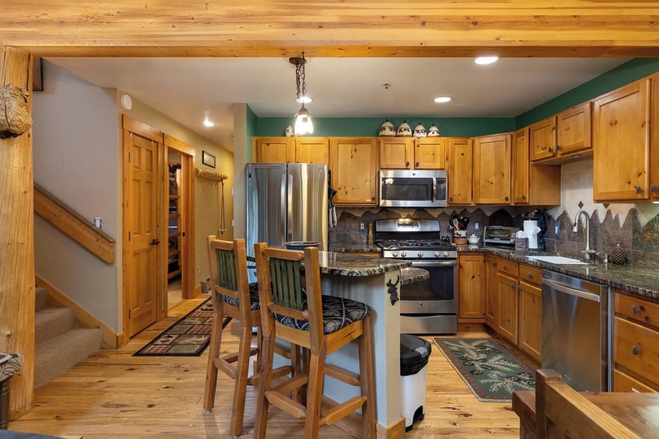 A cozy kitchen with wooden cabinets and a stainless steel stove and refrigerator. A small island with two wooden barstools is in the center. Stairs are adjacent to the kitchen on the left side.