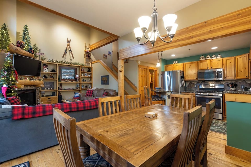 A cozy dining area with a wooden table and chairs is adjacent to a living room with a decorated Christmas tree and fireplace. A kitchen with stainless steel appliances is visible in the background.