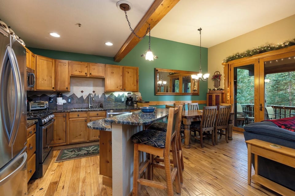 A cozy kitchen and dining area with wooden cabinets, a granite island with stools, a dining table with chairs, wooden floors, and a view of trees outside through a glass door.