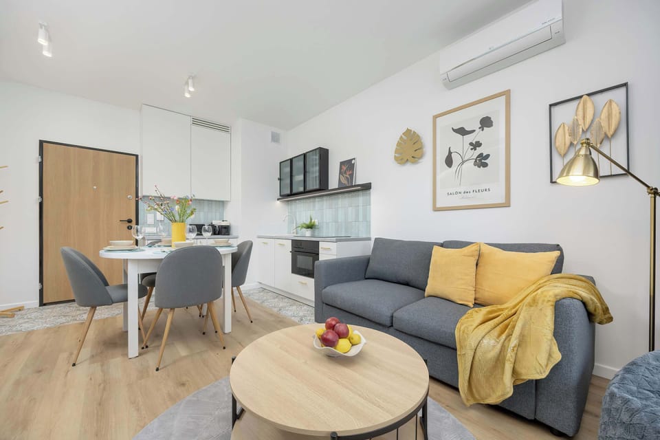 A well-lit living room with a round coffee table, yellow accent cushions, and framed wall art.

