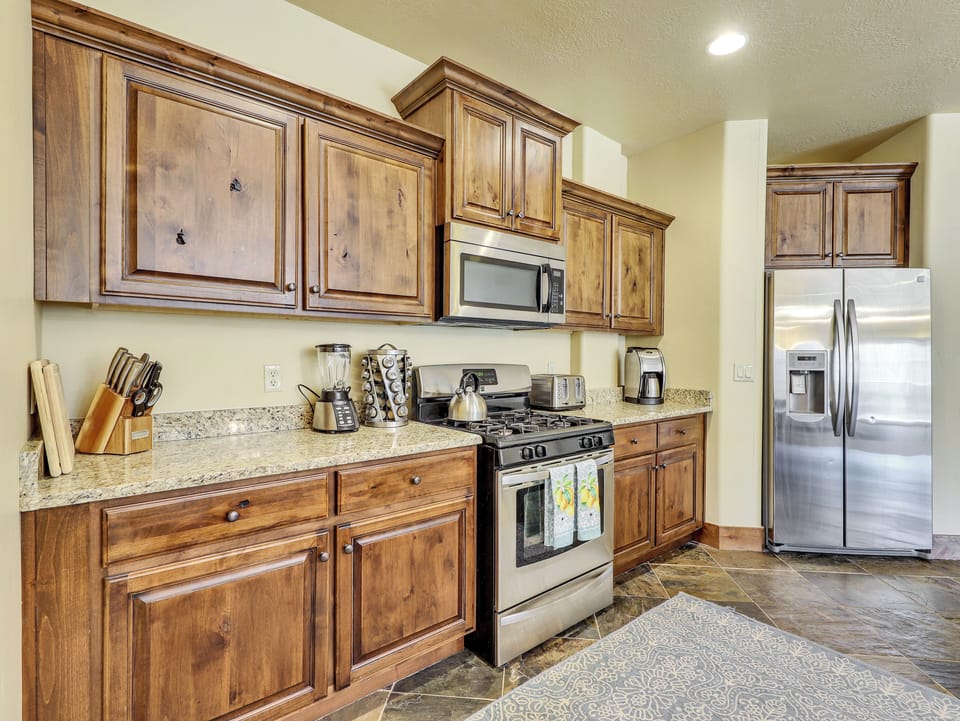 View of fully equipped kitchen featuring a coffee maker, microwave, stove/oven, upgraded cabinetry, granite countertops, blender, kettle, cooking spices, and stainless steel refrigerator