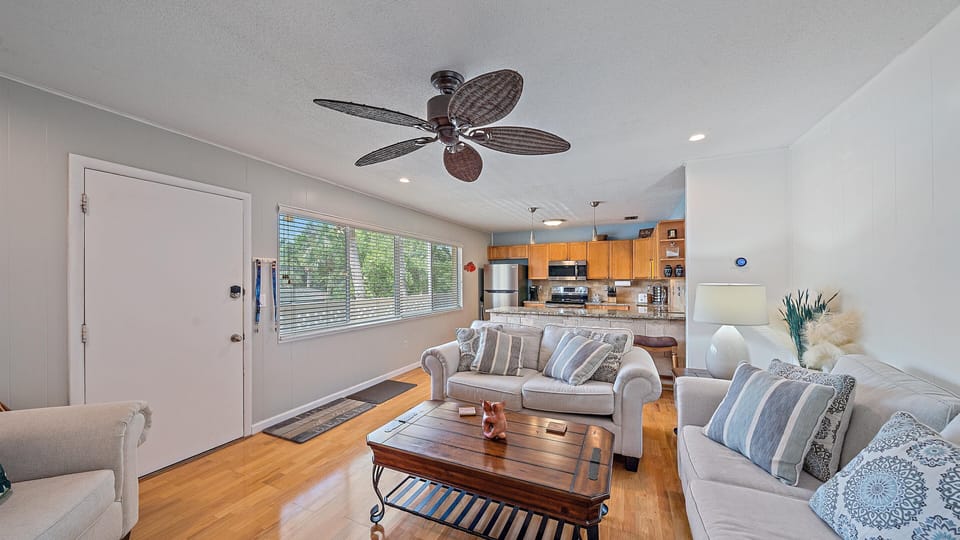 A cozy living room with hardwood floors, light gray walls, and a ceiling fan. It features two upholstered sofas, a wooden coffee table, and an open kitchen with wooden cabinets and modern appliances.