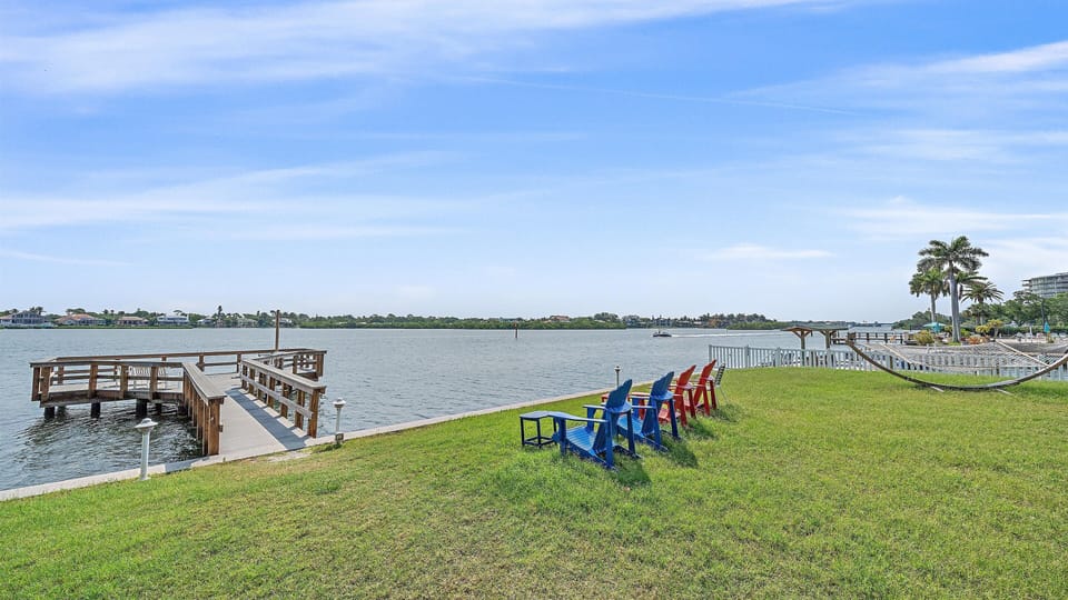 Wooden docks extend into a calm body of water, while colorful Adirondack chairs and a hammock are set on the grassy shore under a clear blue sky.