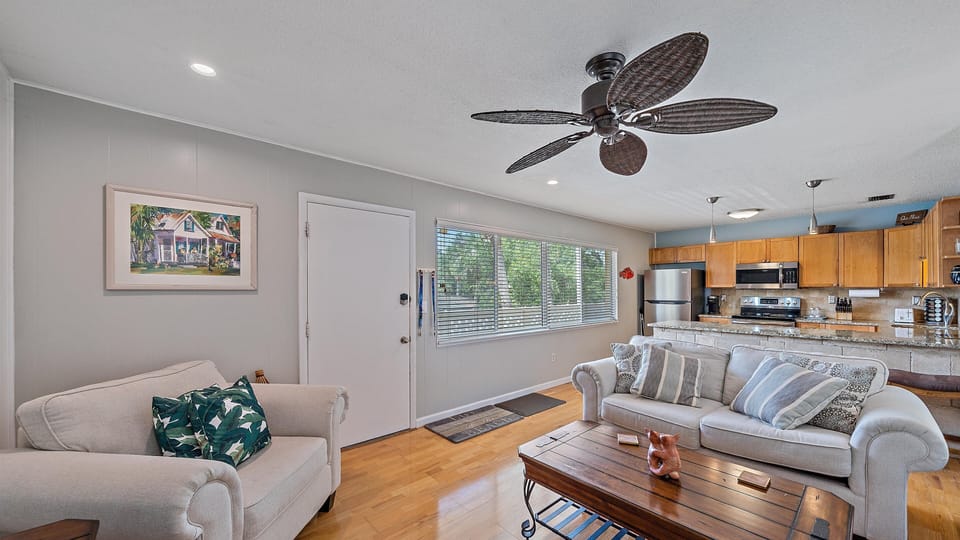 A cozy living room with light-colored walls, armchairs, and a sofa adorned with cushions. A ceiling fan and a painting enhance the space, which opens to a modern kitchen with wooden cabinets.