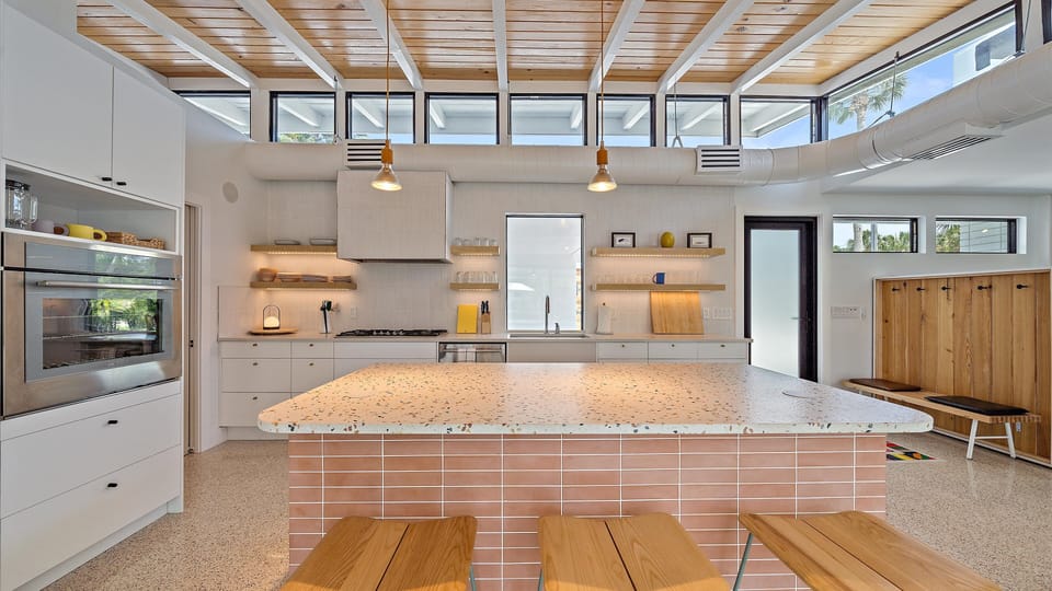 A modern kitchen with a terrazzo island, wooden stools, white cabinetry, built-in appliances, open shelving, and large windows. A row of wooden lockers is visible on the right.