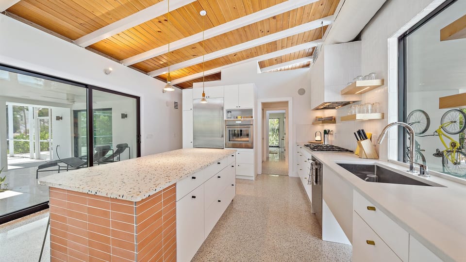 Modern kitchen with white cabinets, a large island with a terrazzo countertop, stainless steel appliances, and wooden ceiling beams. A sliding glass door leads to an enclosed porch area.
