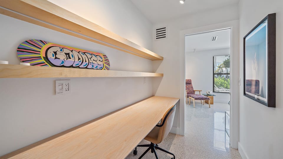 A minimalist home office with a wooden desk, chair, floating shelves, and skateboard decor. An adjacent room with chairs and large windows is visible through the open door.