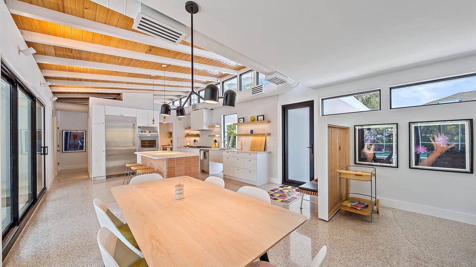 A modern, open-concept kitchen and dining area with white cabinets, wooden accents, pendant lighting, large windows, an island with bar stools, and a dining table with white chairs.